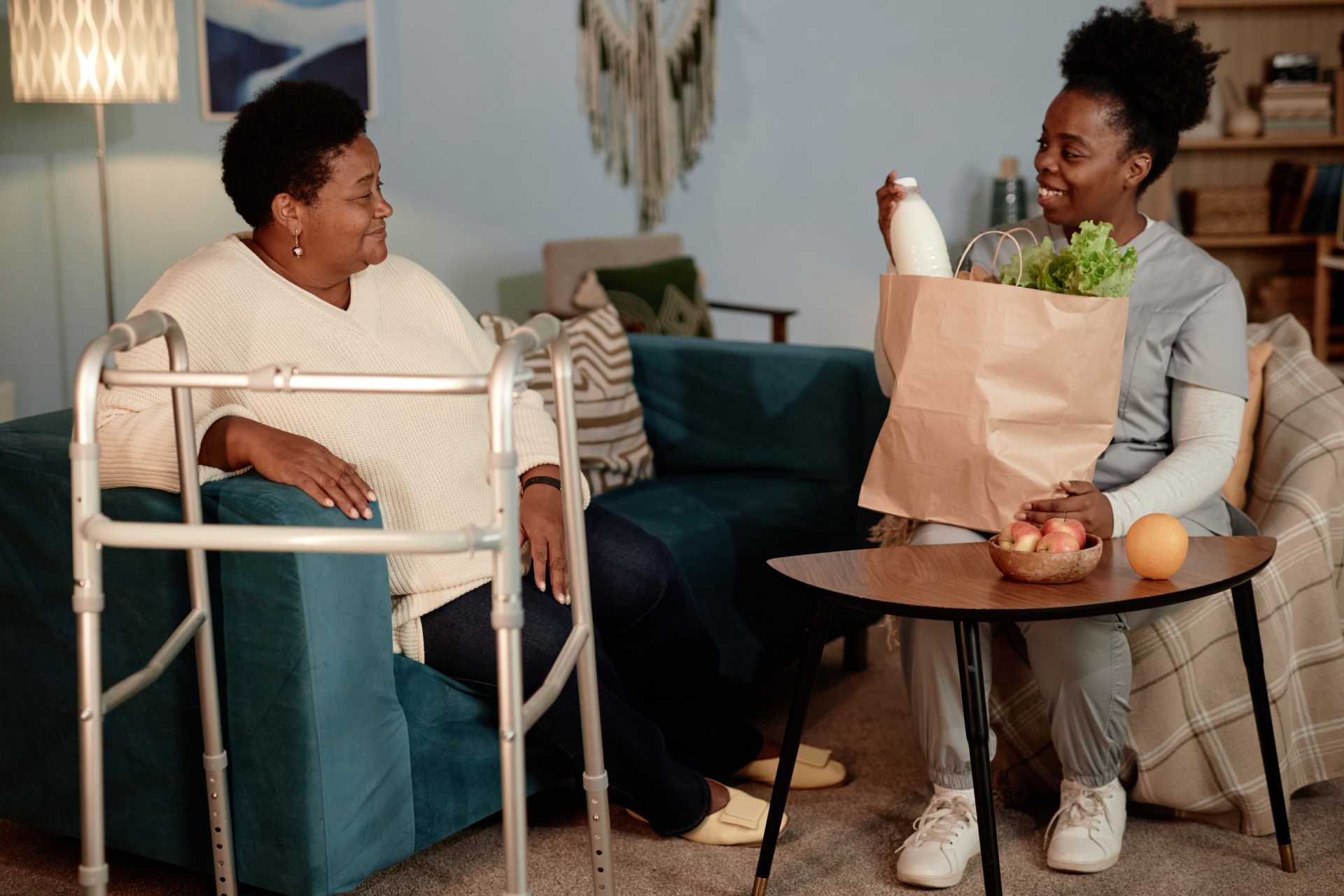 A woman using a walker stands beside another woman seated on a couch, both engaged in conversation