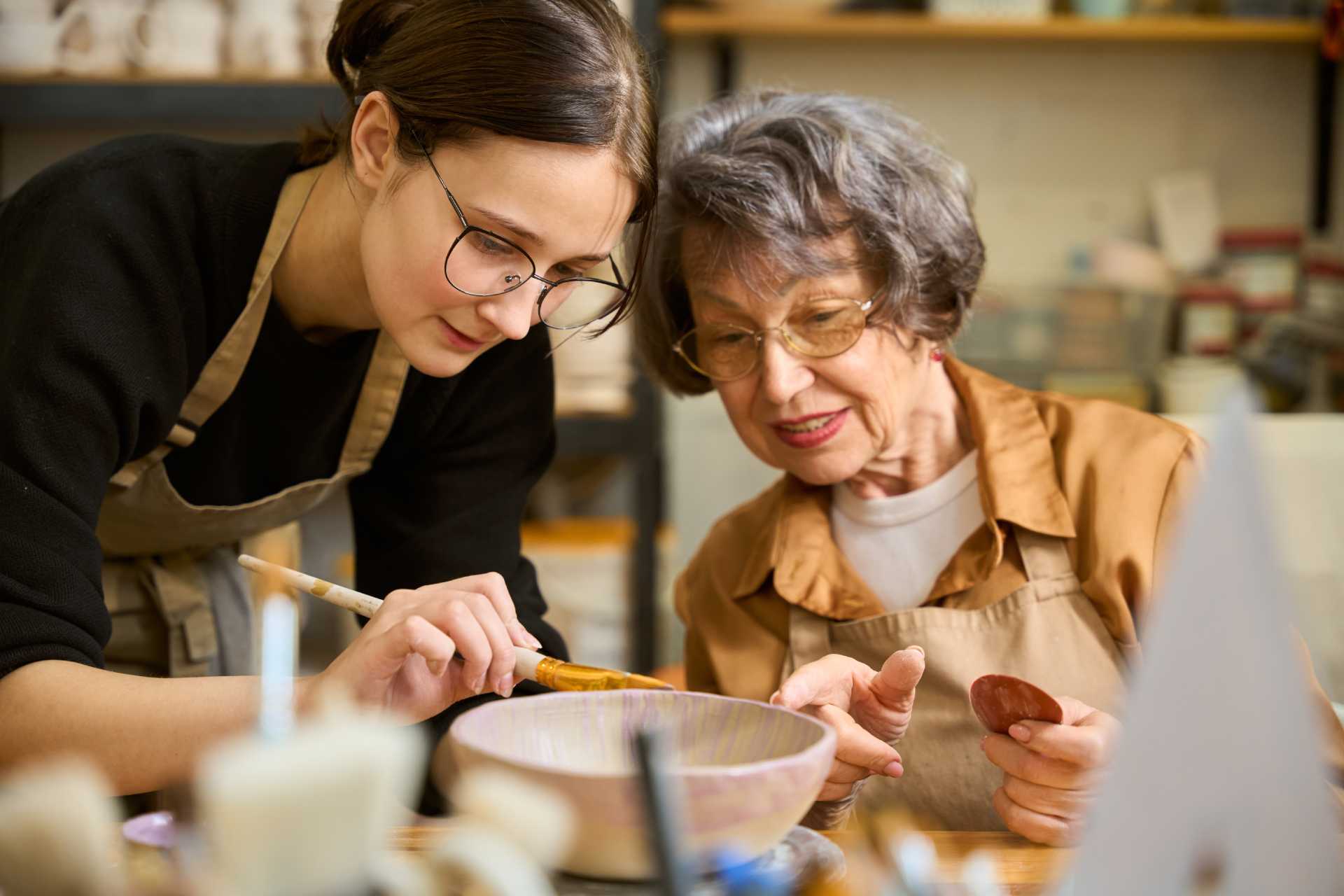 A woman assists an older woman in shaping pottery, demonstrating techniques and providing guidance in a creative setting