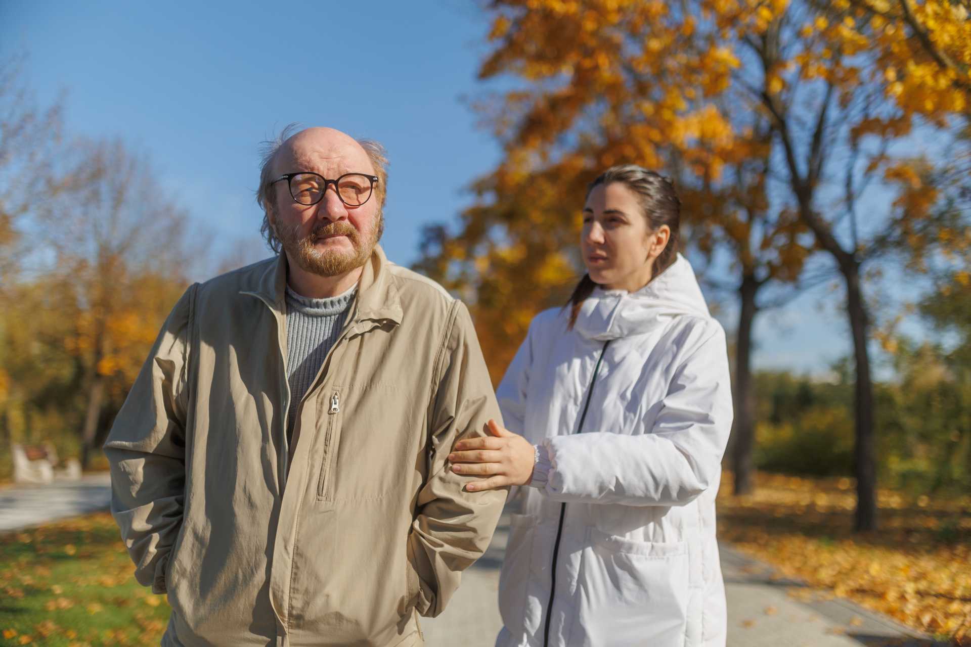 A younger woman in a white coat holds the arm of an older man in a beige jacket as they walk along a park path in autumn