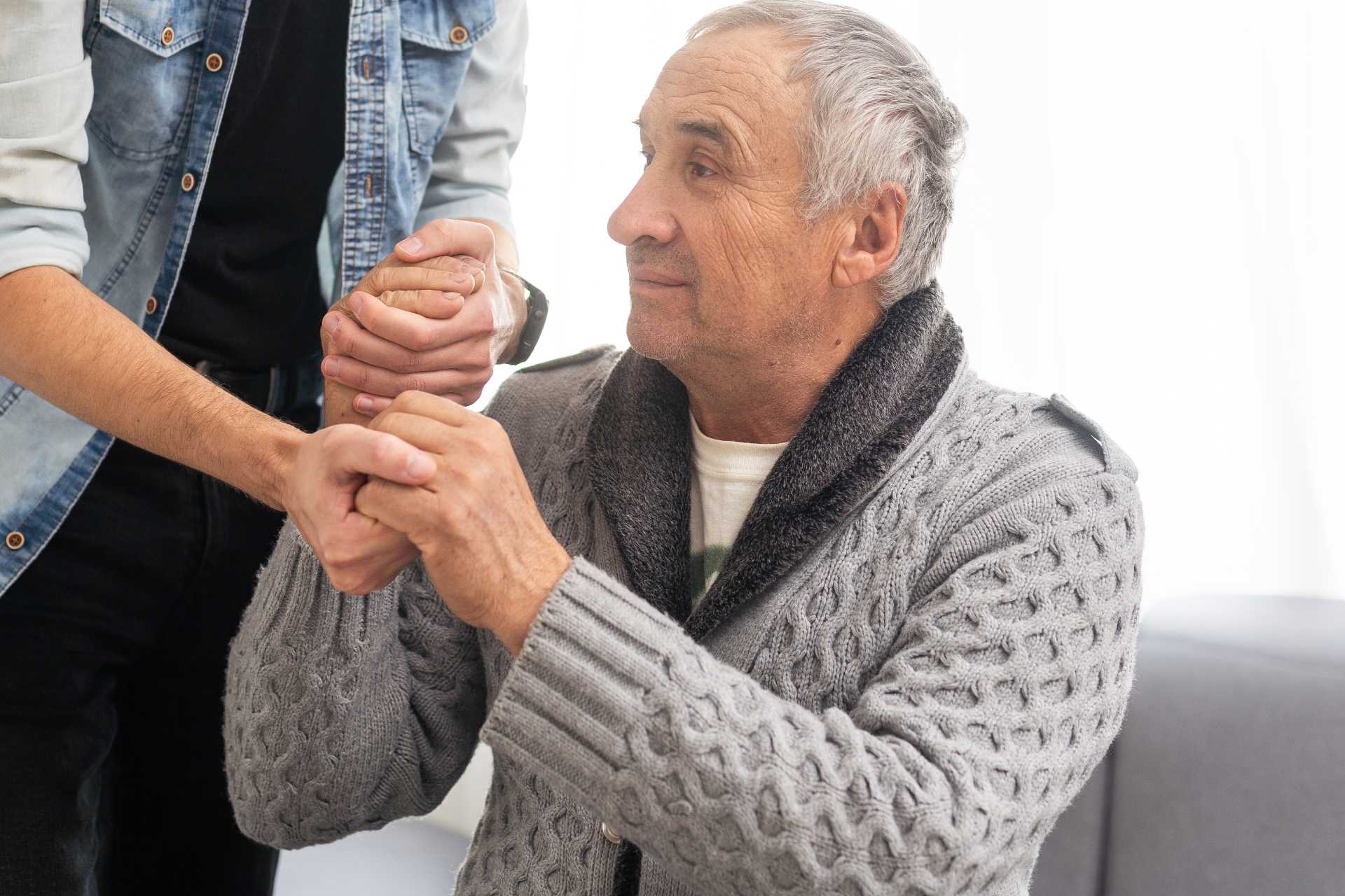 A man gently holds the hand of an older man, conveying a sense of support and connection between them