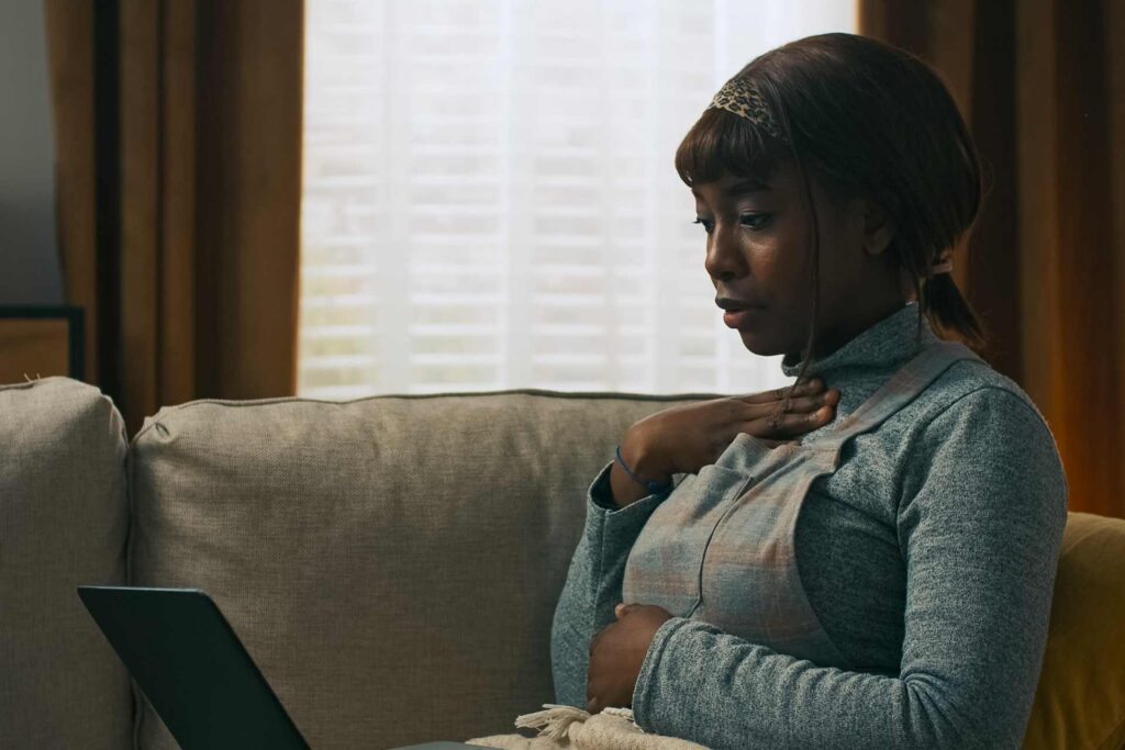 A woman seated on a couch, focused on her laptop, with a cozy living room setting in the background